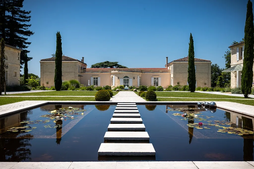 Bordeaux estate featuring a beige-toned mansion with cylindrical towers, symmetrical grounds, a central black and white checkered pathway crossing a reflecting pool with water lilies, and cypress trees flanking the gardens.