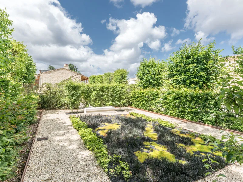 Green rooftop garden with aromatic plants at Château Marquis d'Alesme in Bordeaux, surrounded by lush hedges and historic stone buildings under a cloudy blue sky.