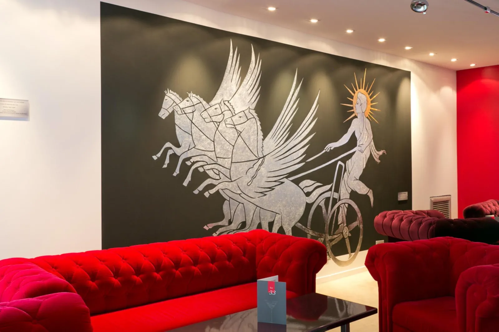 Hotel lounge with red upholstered sofas and a dark wall mural featuring a winged chariot drawn by horses with a divine figure