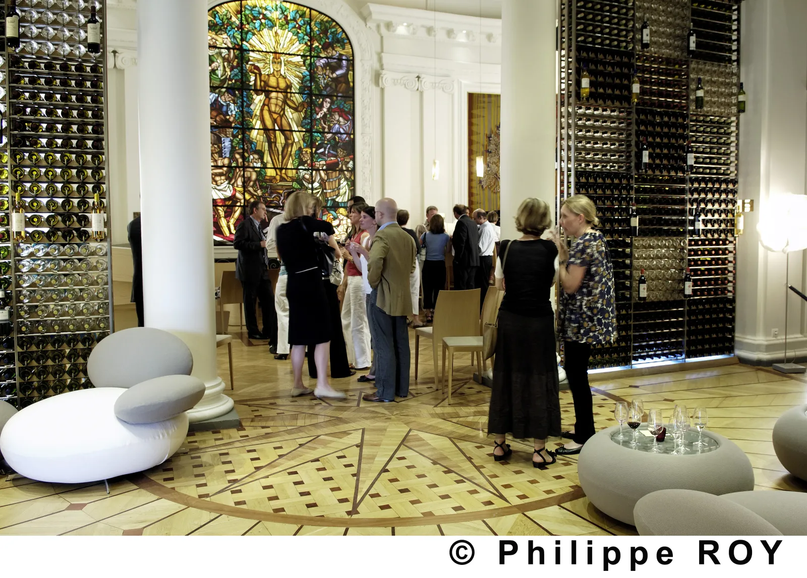 Interior of the CIVB Wine Bar in Bordeaux showing visitors standing in conversation, surrounded by metallic display panels, a colorful stained glass window, and white rounded seating sculptures in the foreground.