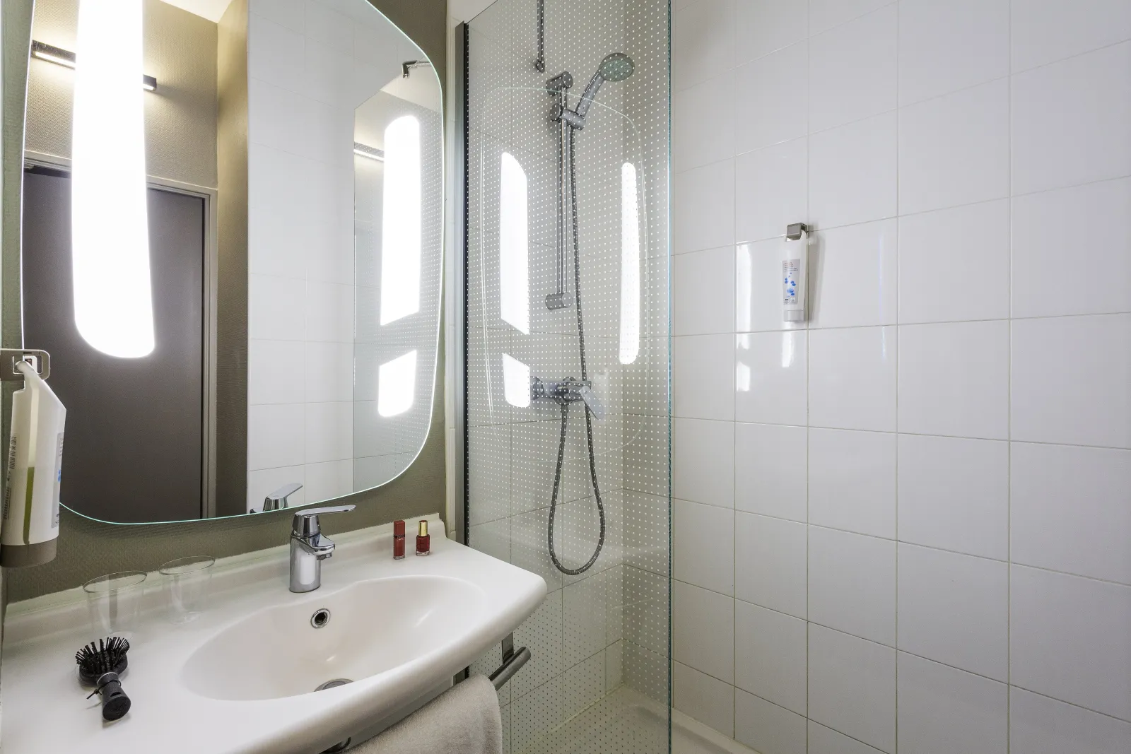 Modern hotel bathroom featuring a white sink, illuminated mirror, walk-in shower with glass partition and light gray tiles