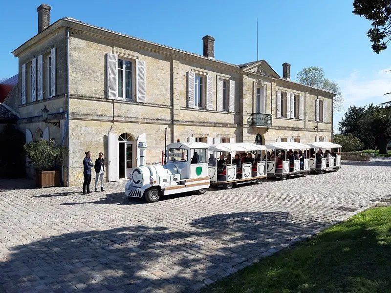 A small white and blue tourist train parked in front of Château Marquis de Vauban in Bordeaux, with passengers aboard and visitors near the stone building's entrance.