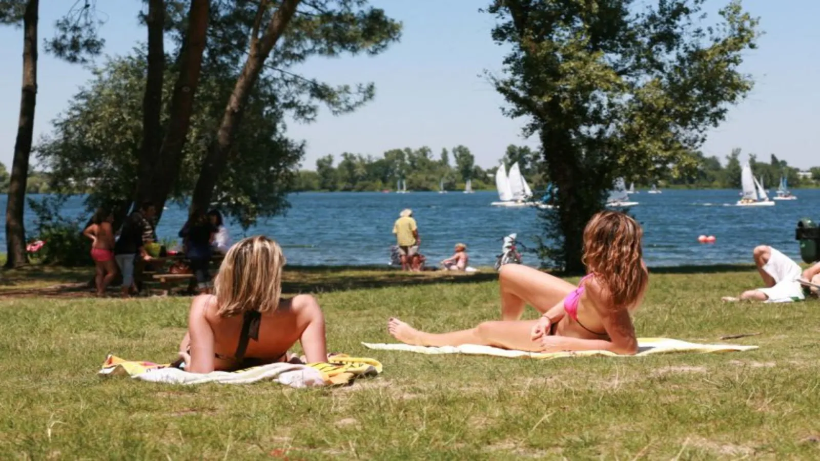 Lake Beach in Bordeaux: visitors relax on the grass overlooking the lake with sailboats. Trees frame the scene while other swimmers enjoy the water in the background.