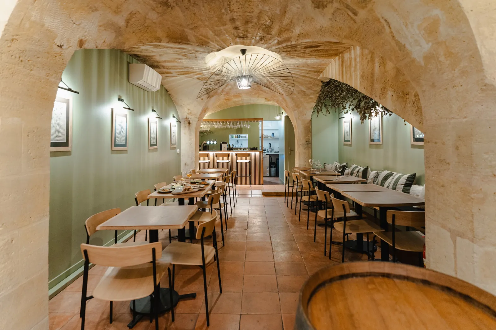 Underground vaulted restaurant interior in Bordeaux featuring wooden tables, modern chairs, pale green walls, exposed stone ceiling and decorative light fixture.