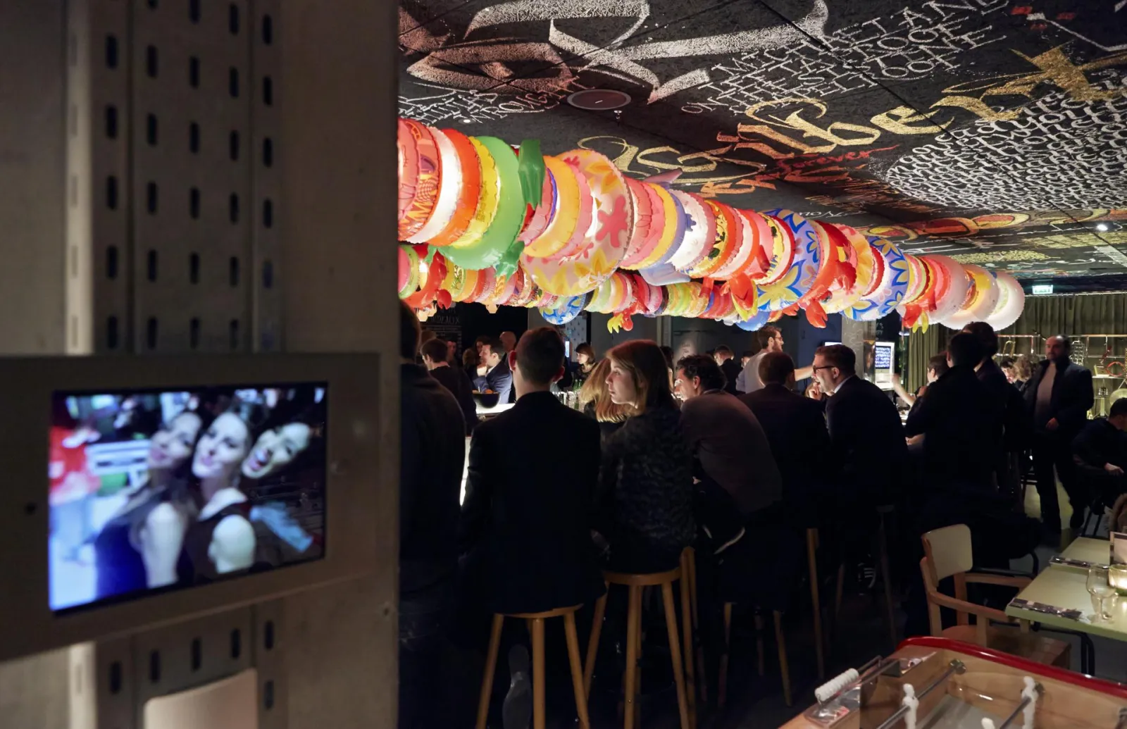 Interior of Mama Shelter Bar in Bordeaux with guests in dark clothing beneath hanging colorful lanterns, with a television screen visible on the left