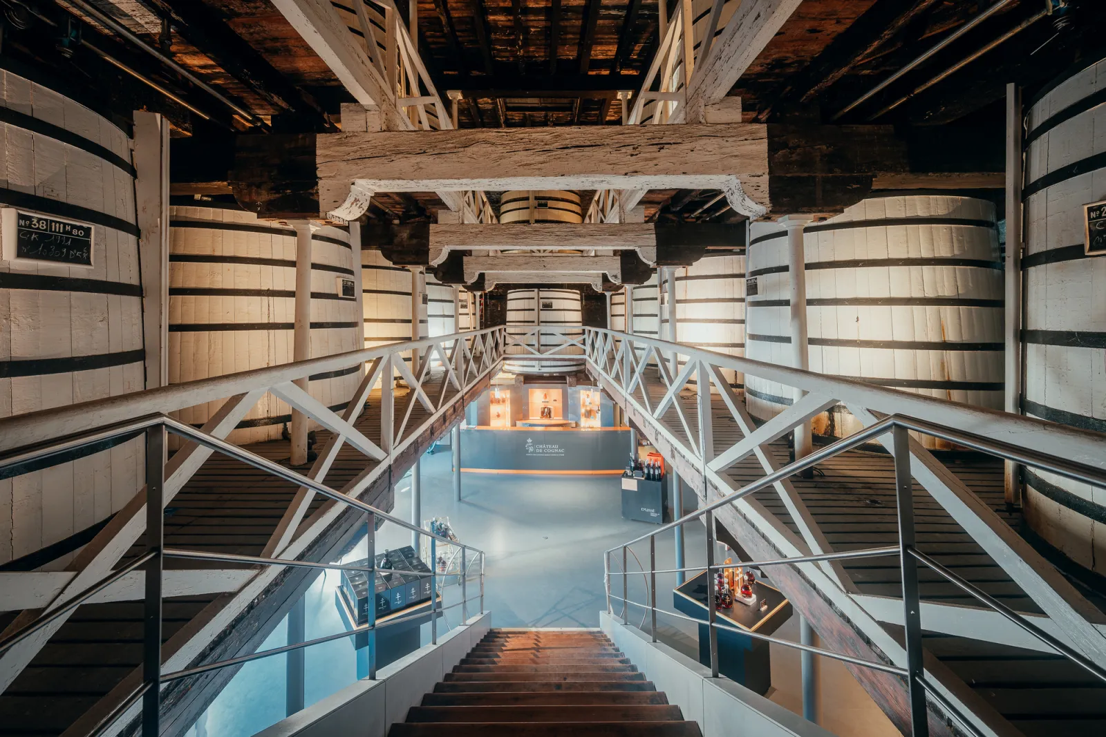 Overhead view of a central staircase descending between two rows of wooden cognac barrels in a historic cellar at Baron Otard Cognac Castle in Bordeaux.
