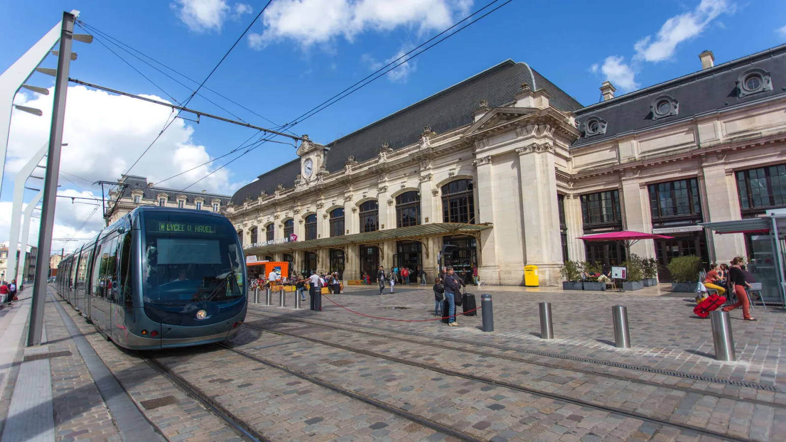 A modern blue tram approaches Bordeaux's Saint-Jean station, with the grand historic cream-colored building featuring ornate architecture and slate roof in the background under a clear blue sky.