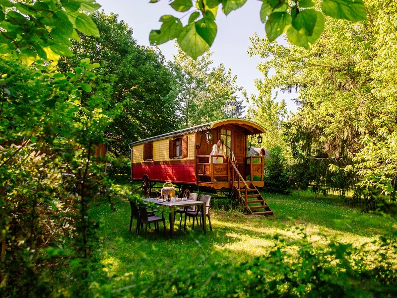 Red and beige wooden caravan surrounded by lush greenery with outdoor dining table at EcÔtelia Estate in Bordeaux