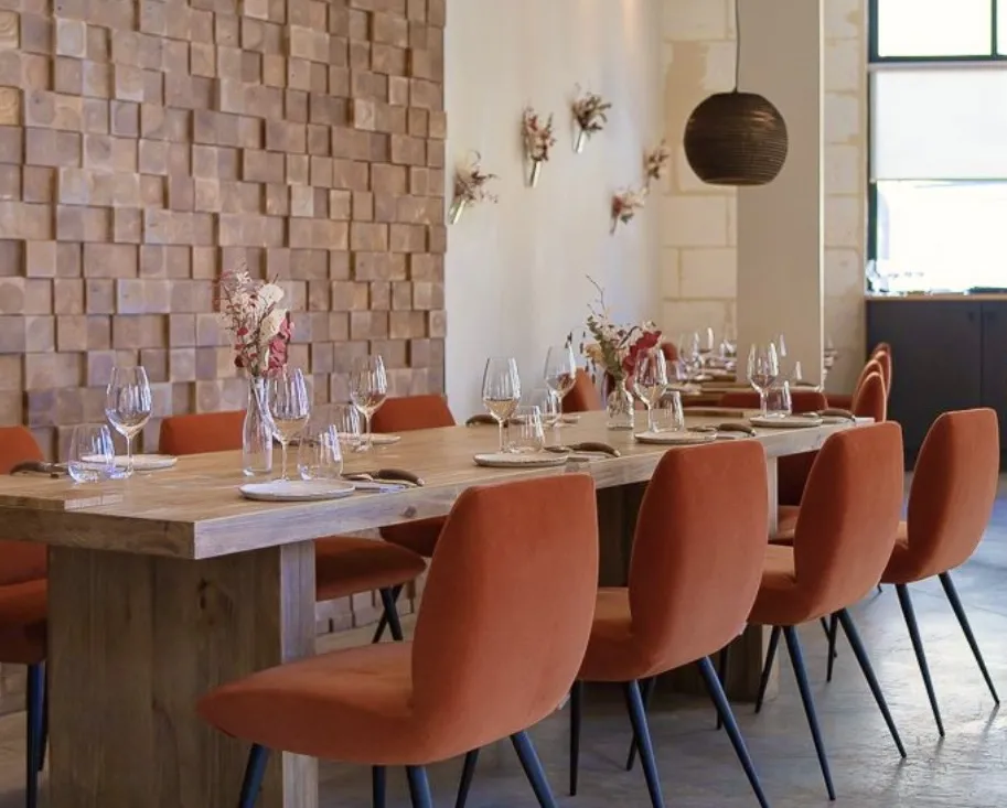 Elegant dining room featuring a light wood table, burnt orange chairs, wine glasses arranged place settings, textured pink brick wall, dried flower wall decor and black pendant light.