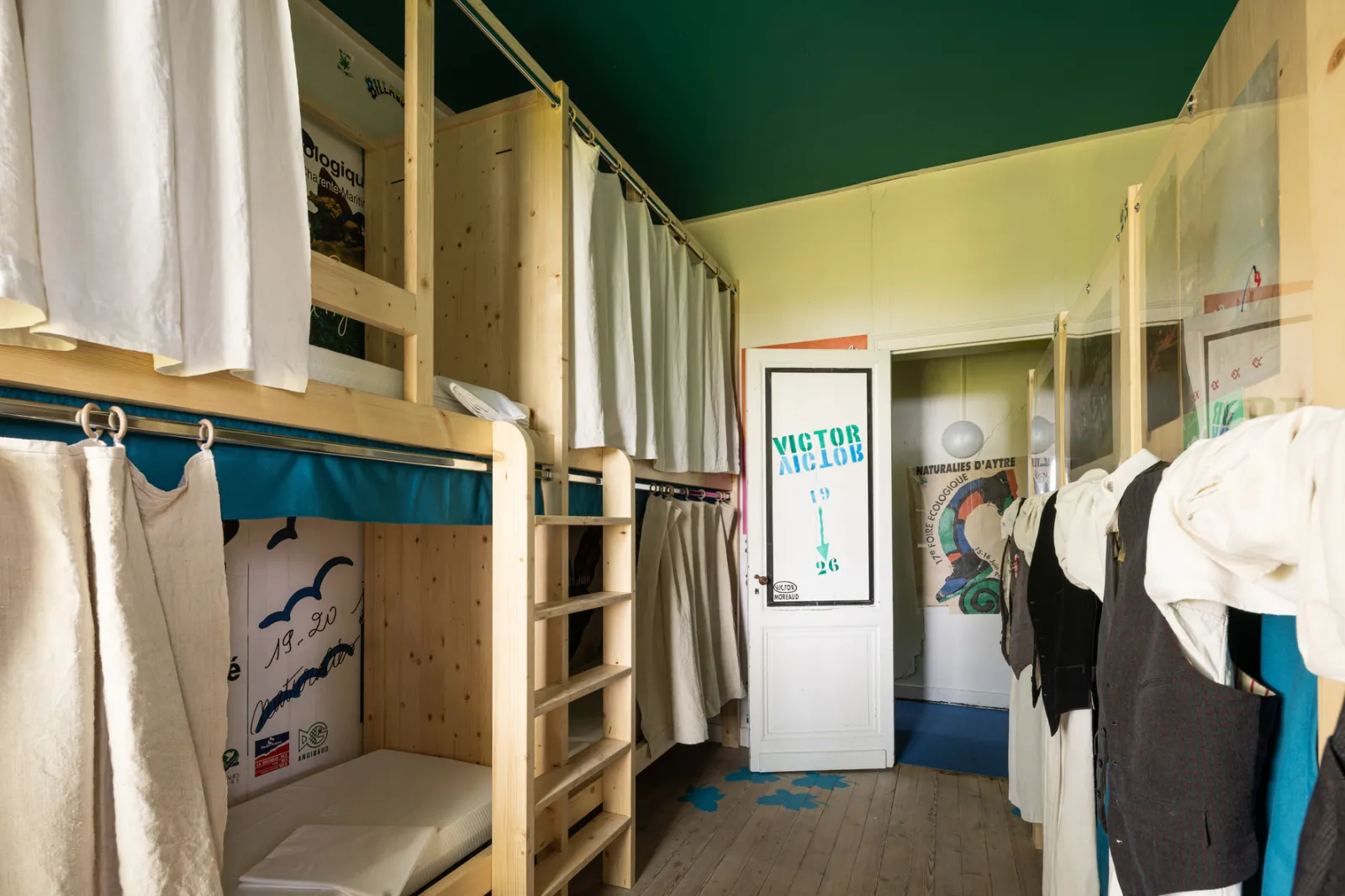Joualles dormitory in Bordeaux featuring wooden bunk beds with white and gray curtains, a Victor Arlaud poster, and clothes hanging on the beds