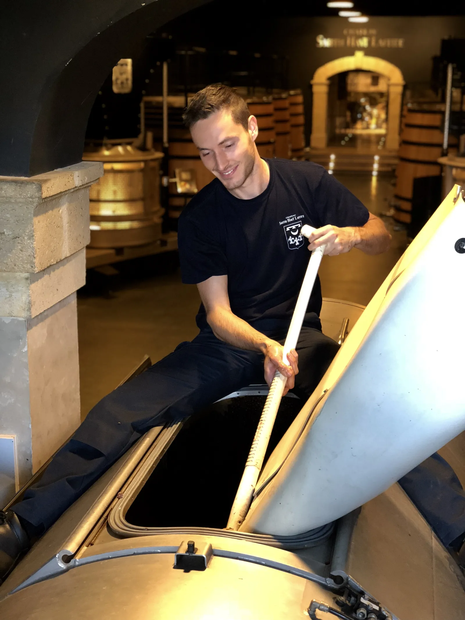 A man in a black t-shirt handles a glass tube in a wine production cellar at Château Smith Haut Lafitte in Bordeaux, with golden barrels visible in the background.