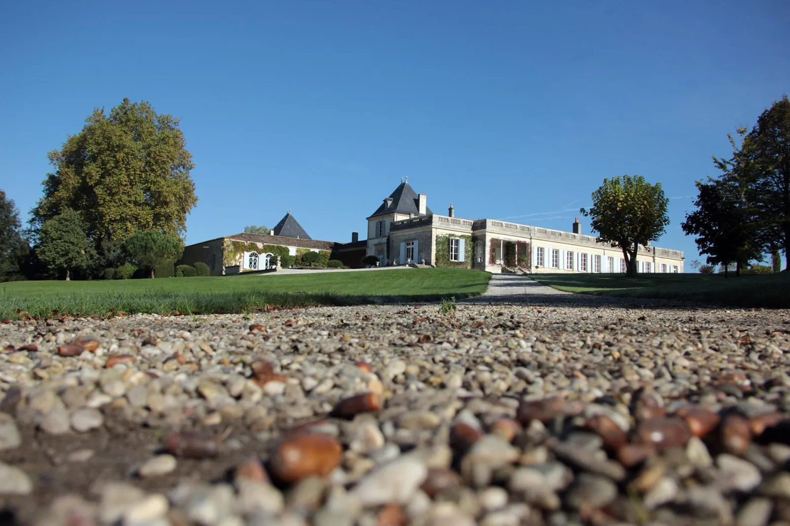 View of Château Paveil de Luze near Bordeaux, a wine estate featuring white stone buildings with mansard roofs, surrounded by manicured green lawns and trees, photographed from a gravel driveway.