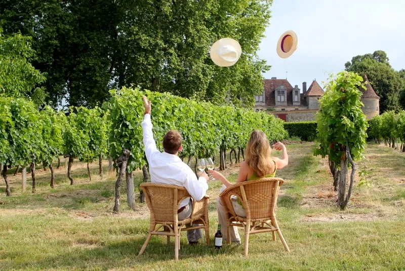 Two people sitting in wicker chairs in a vineyard, tossing their hats in the air. A historic castle is visible in the background.