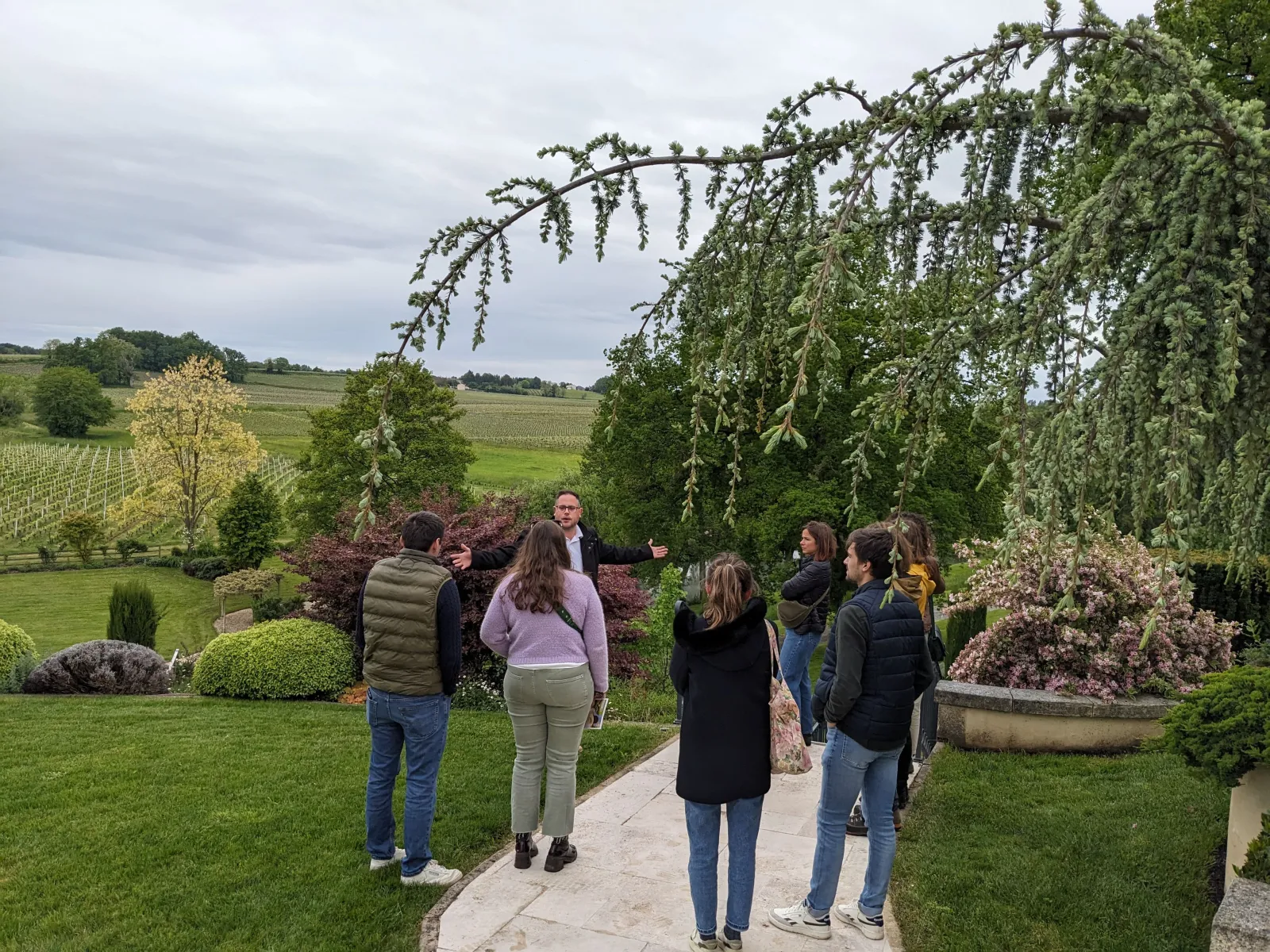 A group of visitors on a guided tour in the gardens of Château La Rose Perrière near Bordeaux, with views of surrounding vineyards and a weeping willow in the foreground.