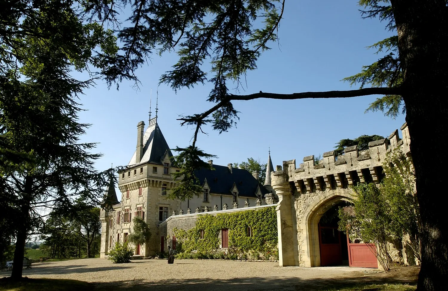 Château de Pressac in Bordeaux, a medieval stone fortress featuring crenellated towers, pointed roof, and red entrance gate, framed by ancient trees.