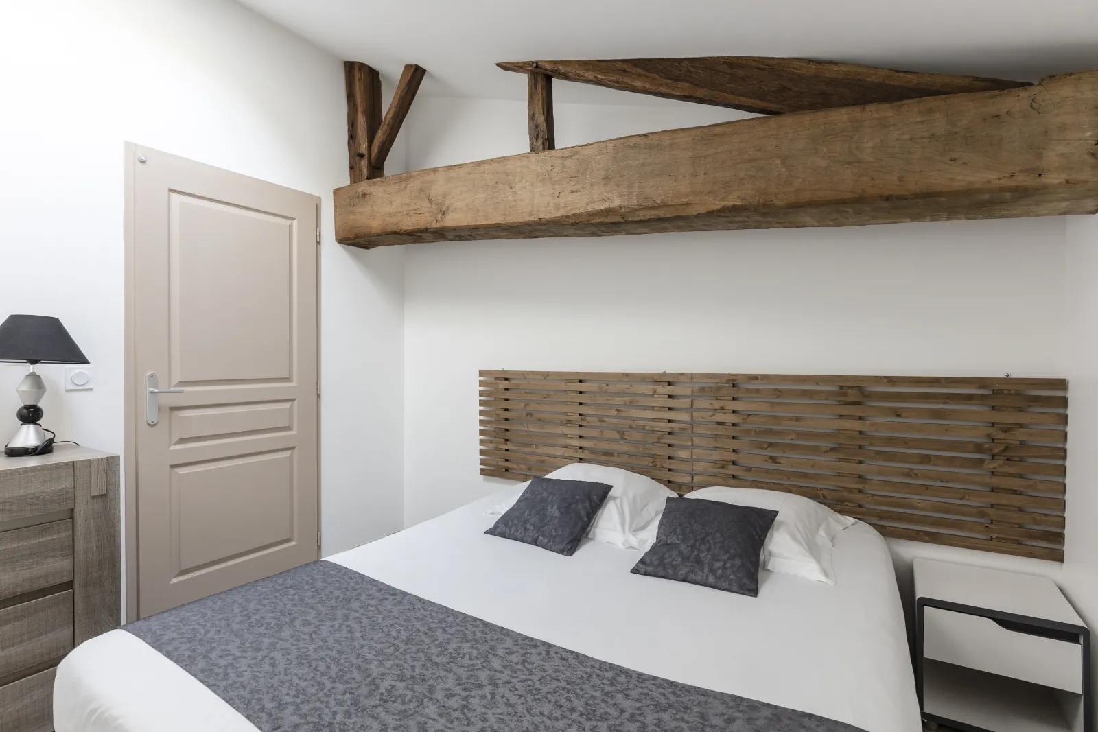 Studio bedroom with double bed, white linens and gray pillows, slatted wooden headboard, exposed ceiling beams, beige door and light wood furniture at Château Pierre de Montignac in Bordeaux