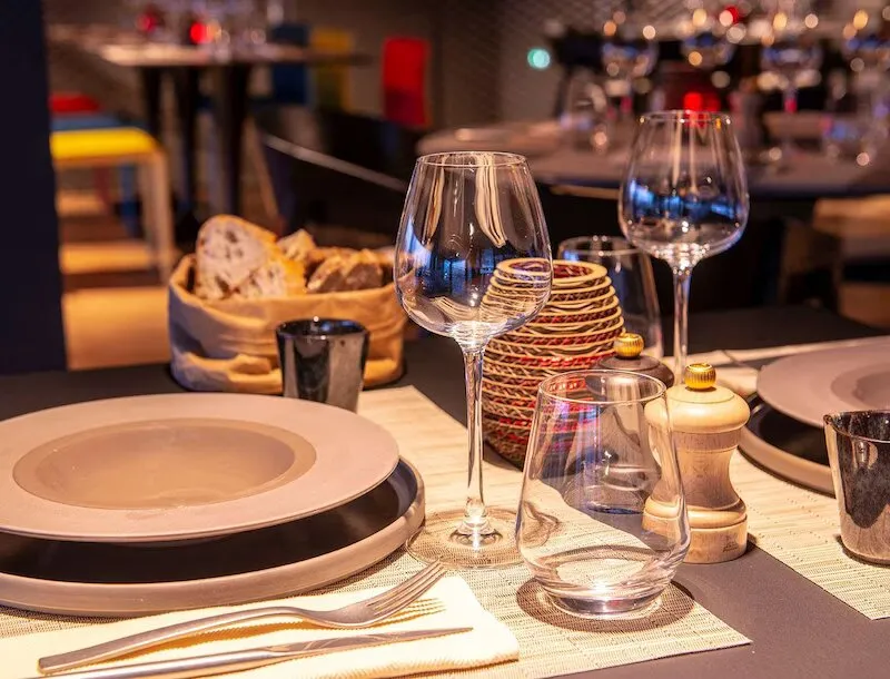 Elegantly set dining table at Wellman Restaurant & Bar in Bordeaux featuring white plates, water glasses, gold cutlery, napkins, and a bread basket in the background, with warm sophisticated lighting.