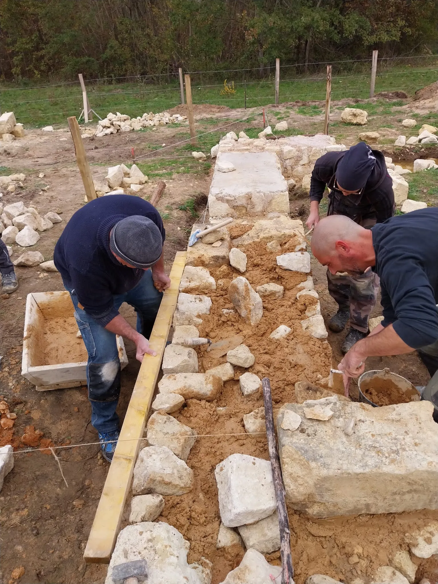 Three workers build a dressed stone wall on a medieval reconstruction site, laying white limestone blocks with mortar in foundation trenches.
