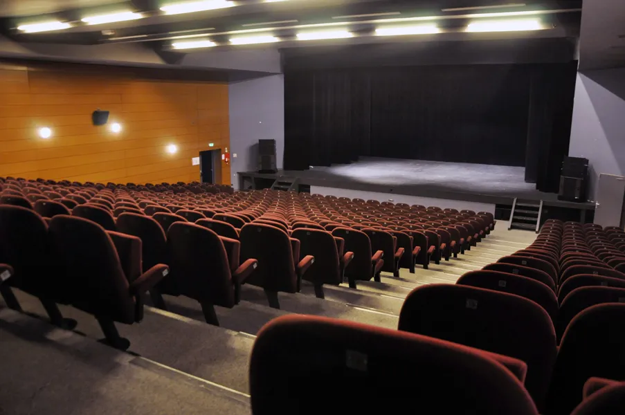 Interior of a cinema hall with empty rows of red seats facing an illuminated stage, with orange and gray walls, at L'Entrepôt du Haillan in Bordeaux.