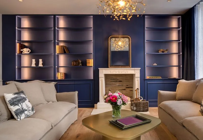 Elegant living room with navy blue walls, illuminated built-in bookshelves, central fireplace, two beige sofas facing each other, gold coffee table, glass chandelier and fresh flowers