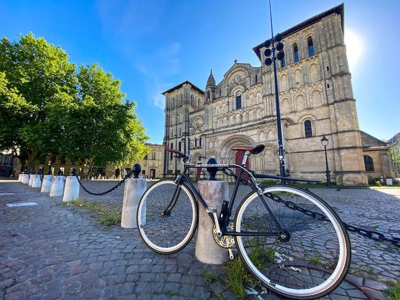Black and white bicycle parked on a cobblestone square in front of the Basilica of Saint-Seurin in Bordeaux, with green trees and blue sky