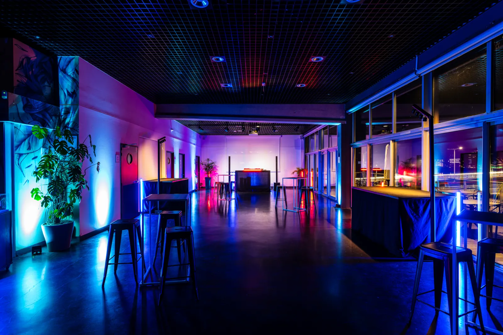 Modern event space interior in Bordeaux featuring blue, pink and purple LED lighting, high-top tables, black bar stools, decorative plants, and floor-to-ceiling windows overlooking the night cityscape.