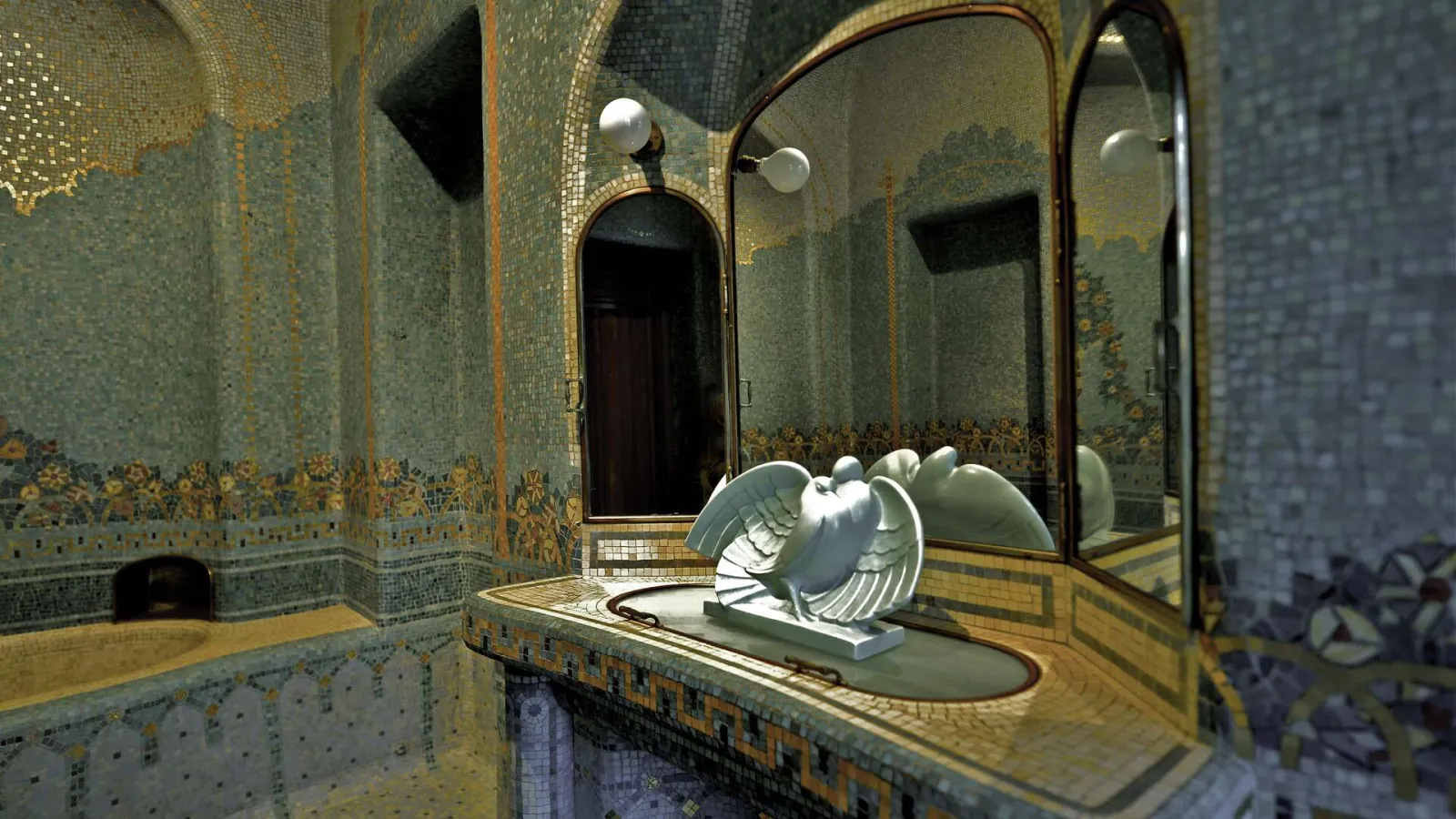Luxurious bathroom of the Frugès Hotel in Bordeaux featuring gold mosaic tiles, arched mirrors, wall sconces, and white angel sculpture on the sink