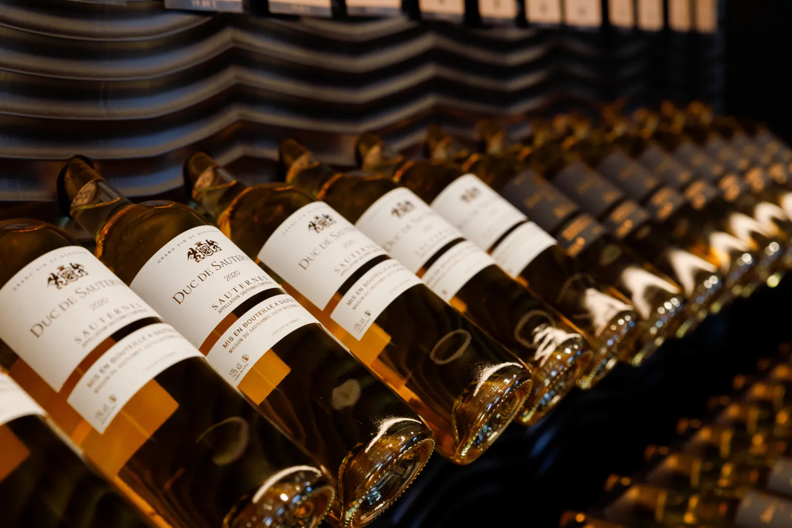 Row of Sauternes bottles with white « Duc de Sauternes » labels displayed on wooden shelves, showcasing the characteristic golden wine from Bordeaux region.
