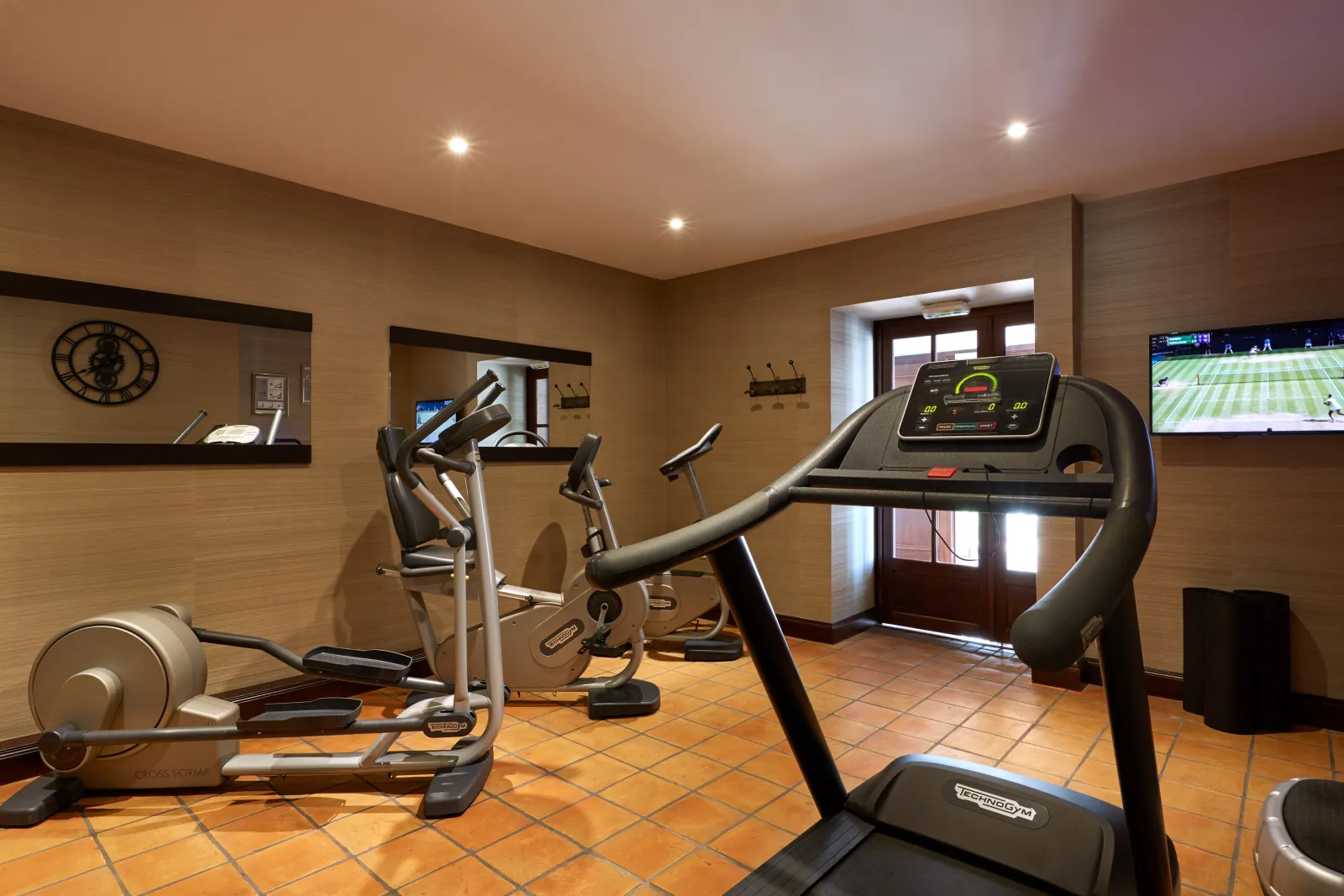 Modern fitness room at Spa Grand Barrail in Bordeaux featuring treadmills, elliptical bikes, television screens, and wall clock, with terracotta flooring and minimalist décor.