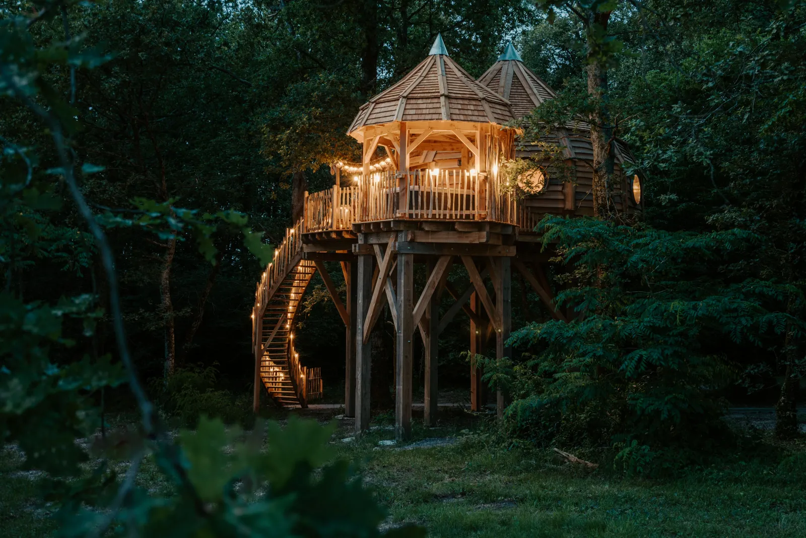 Illuminated wooden treehouse structure at night featuring conical roofs and turrets, elevated on wooden pillars surrounded by dense forest vegetation.