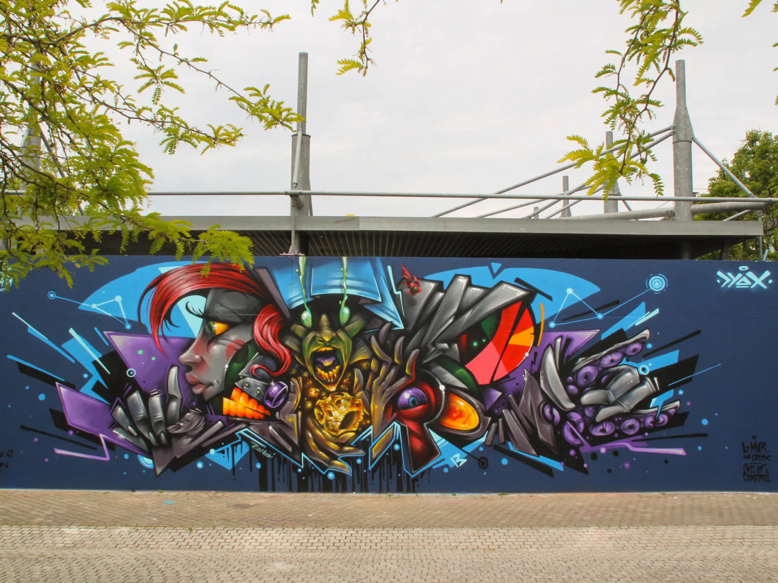 Colorful street art mural on the M.U.R wall in Bordeaux, featuring stylized faces with geometric shapes in blue, red, gold, and purple tones on a dark blue background, beneath a modern bridge.