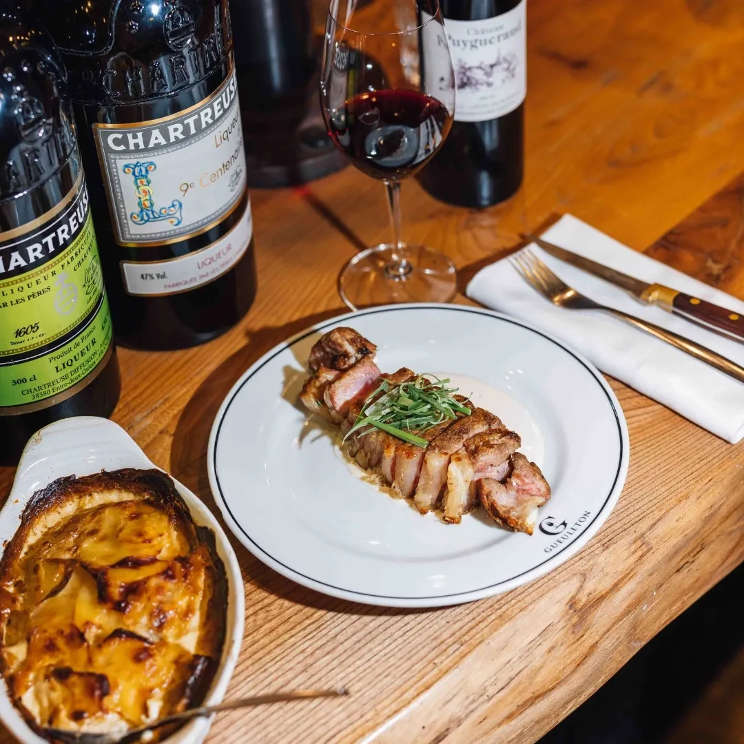 Sliced duck breast plated with vegetables, accompanied by sweet potato puree and Chartreuse Bordeaux wine bottles in a fine dining restaurant setting.
