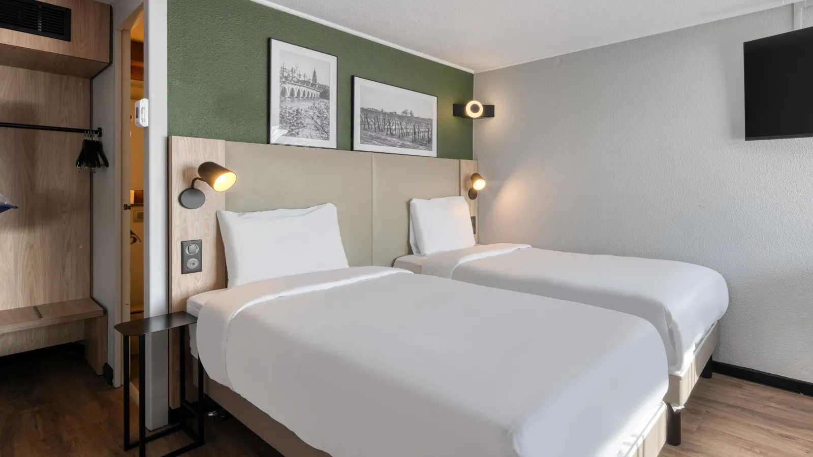 Modern hotel room featuring two white twin beds, beige headboard, sage green accent wall, black and white architectural photographs, contemporary lighting, and light wood furniture