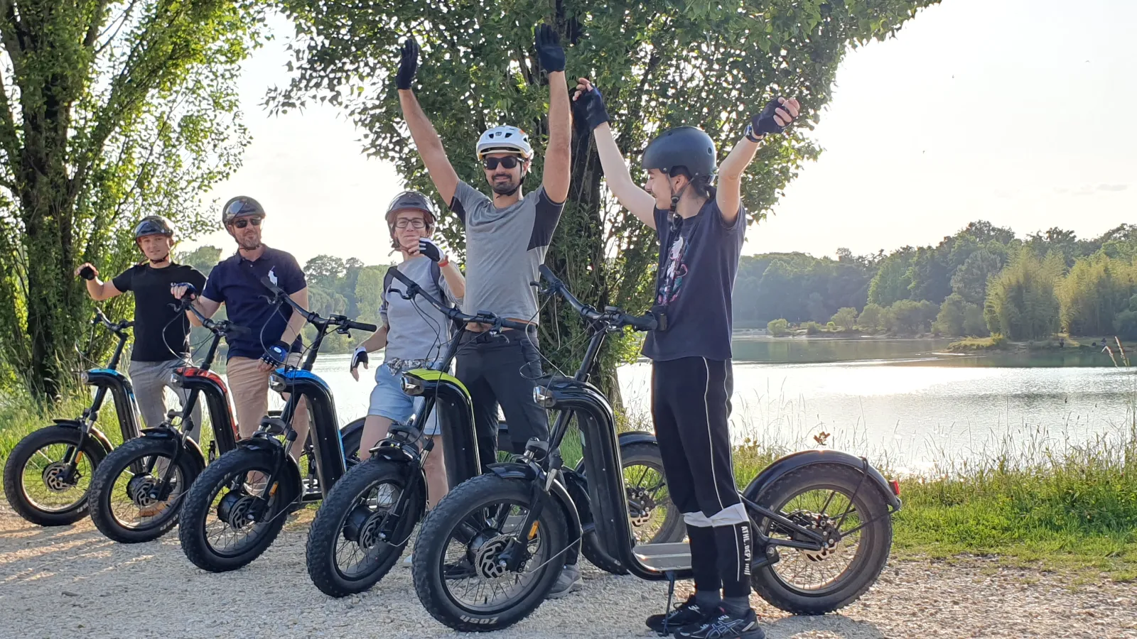 Group of six people on bikes and electric scooters with arms raised, posing on a riverbank path in Bordeaux surrounded by green vegetation.