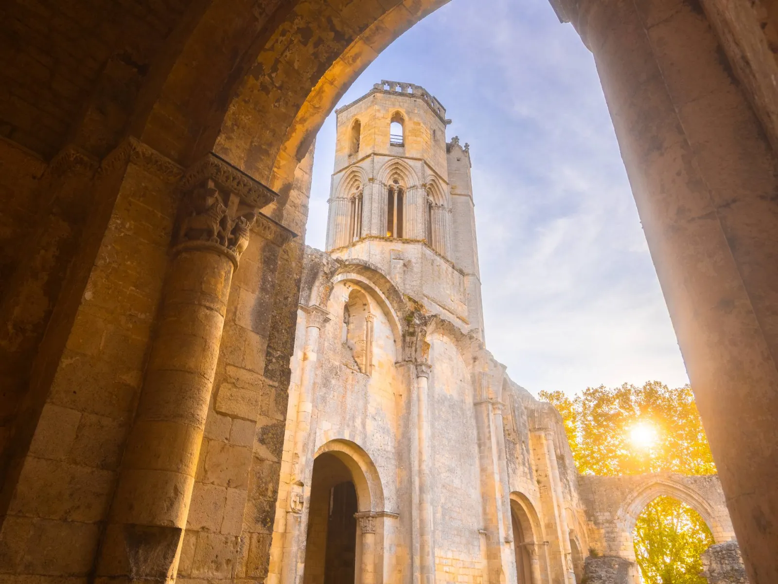 View of the Abbey of La Sauve-Majeure in Bordeaux through a stone archway, showing the Romanesque tower with Gothic windows against a blue sky.