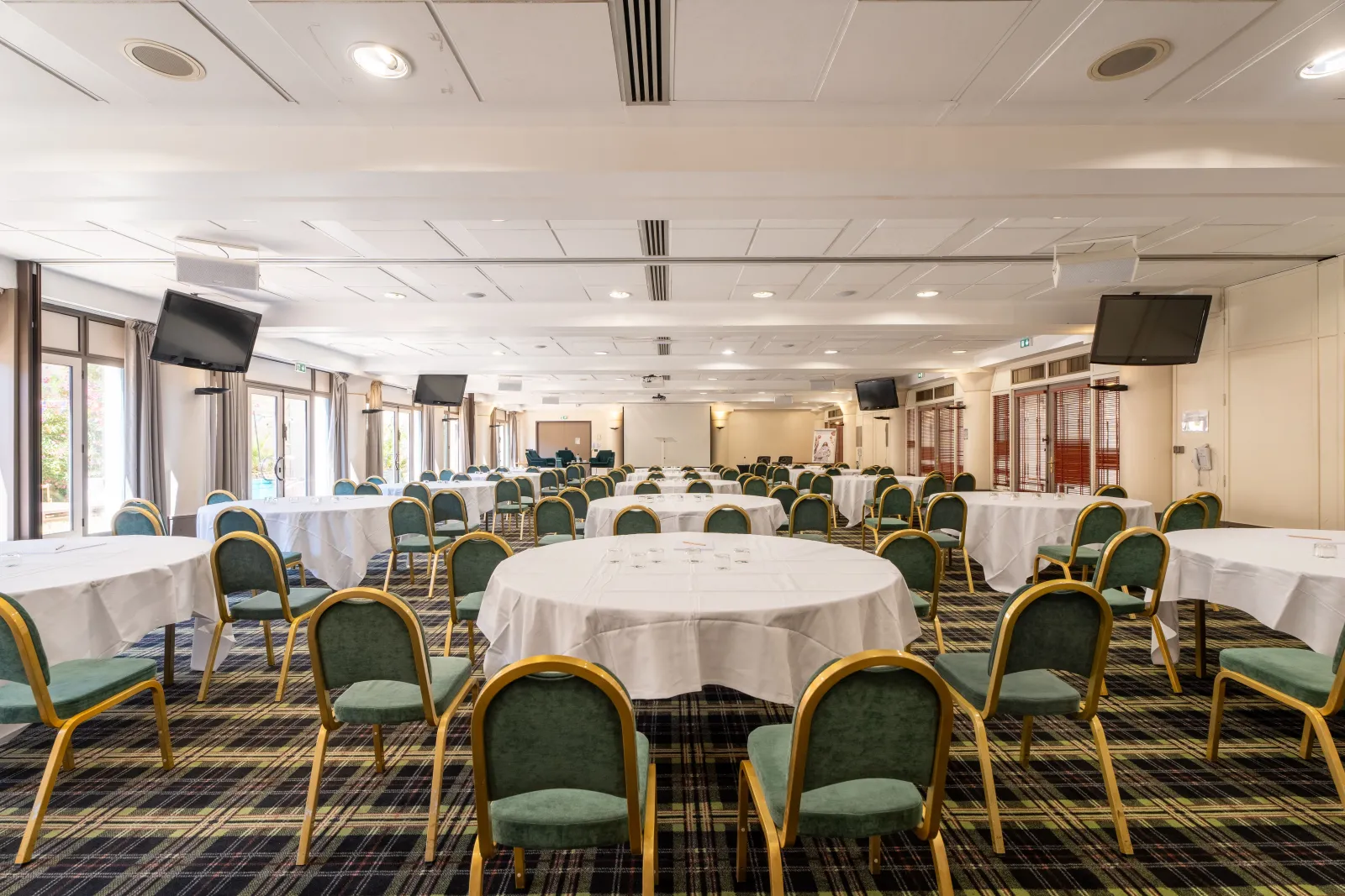 Large conference room at Mercure Bordeaux Château Chartrons featuring white round tables, green and gold chairs, wall-mounted screens, and modern ceiling.