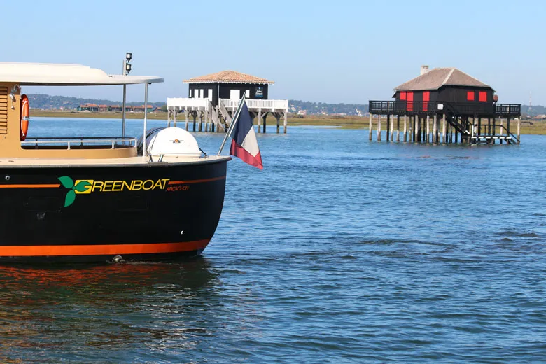 A black and orange boat named Greenboat in the foreground with the French flag, facing two traditional wooden cabins on stilts in Arcachon Bay.