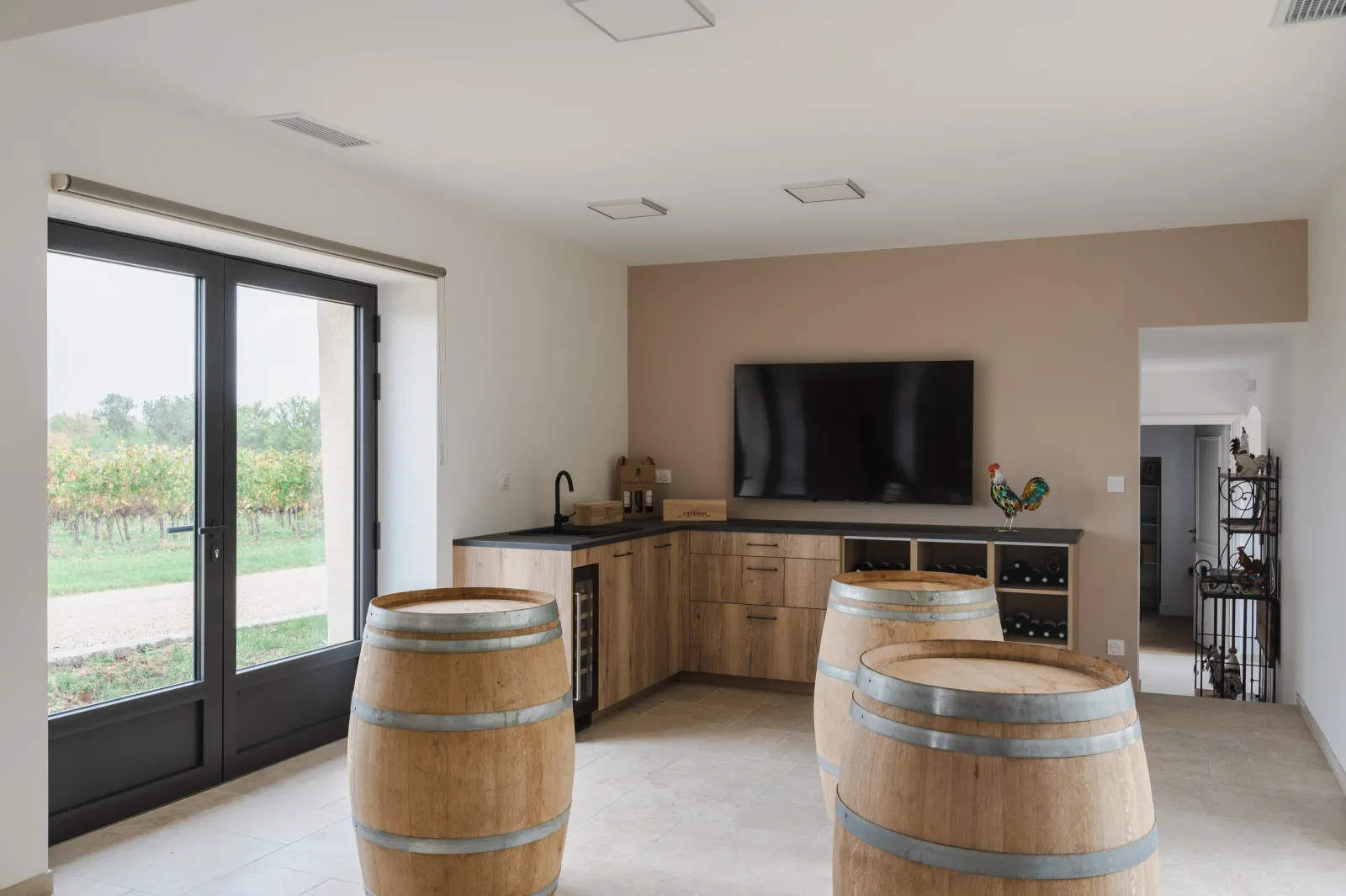 Modern tasting room at Château l'Éperon in Bordeaux, featuring two wooden oak barrels in the foreground, wooden cabinetry, a wall-mounted television, and large glass doors overlooking the vineyards