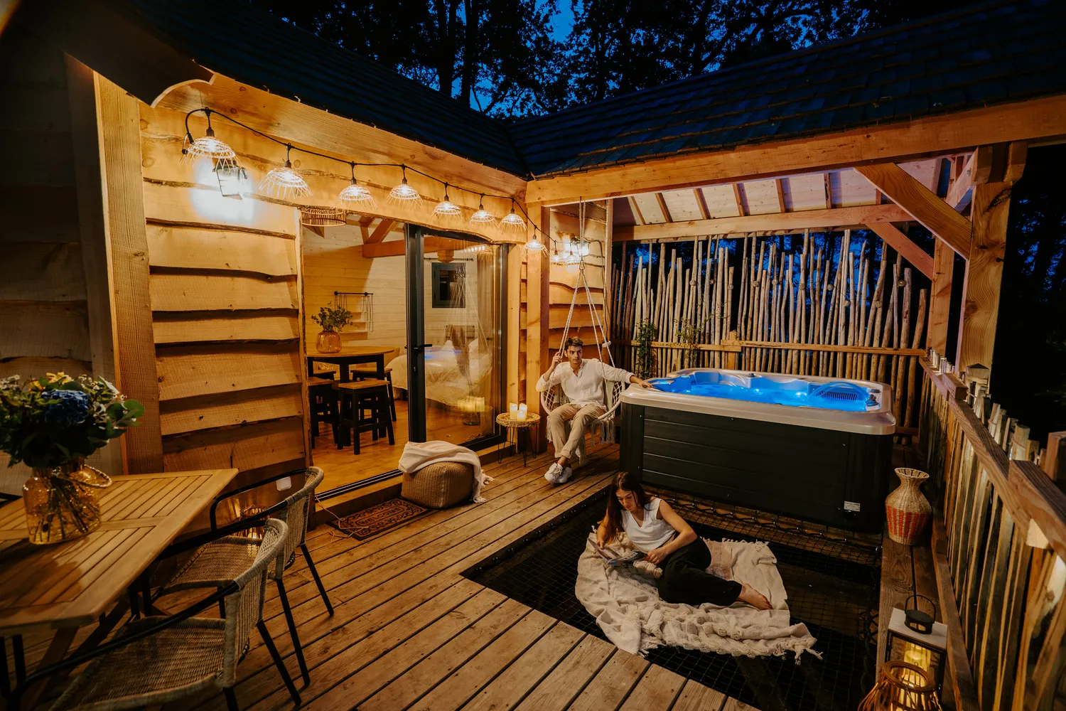 A wooden Girondine cabin with covered terrace, illuminated blue hot tub, two guests relaxing, lounge furniture and string lights at night in Bordeaux