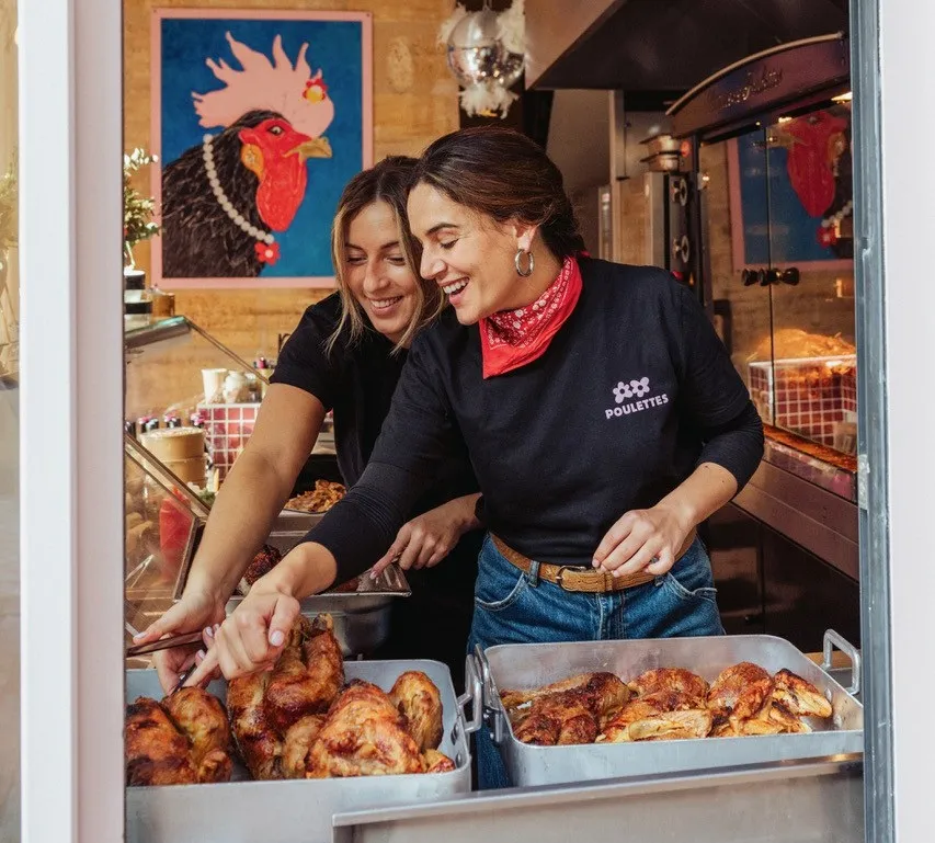 Two smiling women preparing roasted chicken in the display window of Rôtisserie Poulettes in Bordeaux, with a colorful rooster painting in the background.
