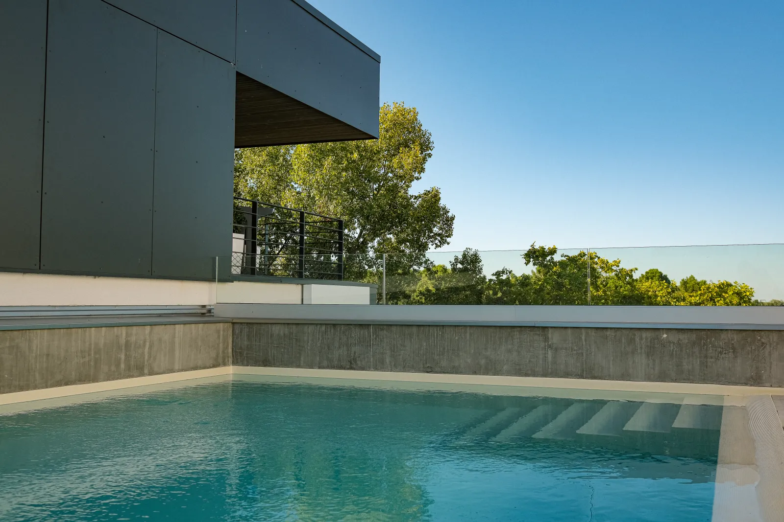 Modern outdoor swimming pool at Ibis Styles Bordeaux Lac Bruges hotel featuring contemporary architecture, concrete terrace and views of surrounding trees under clear sky