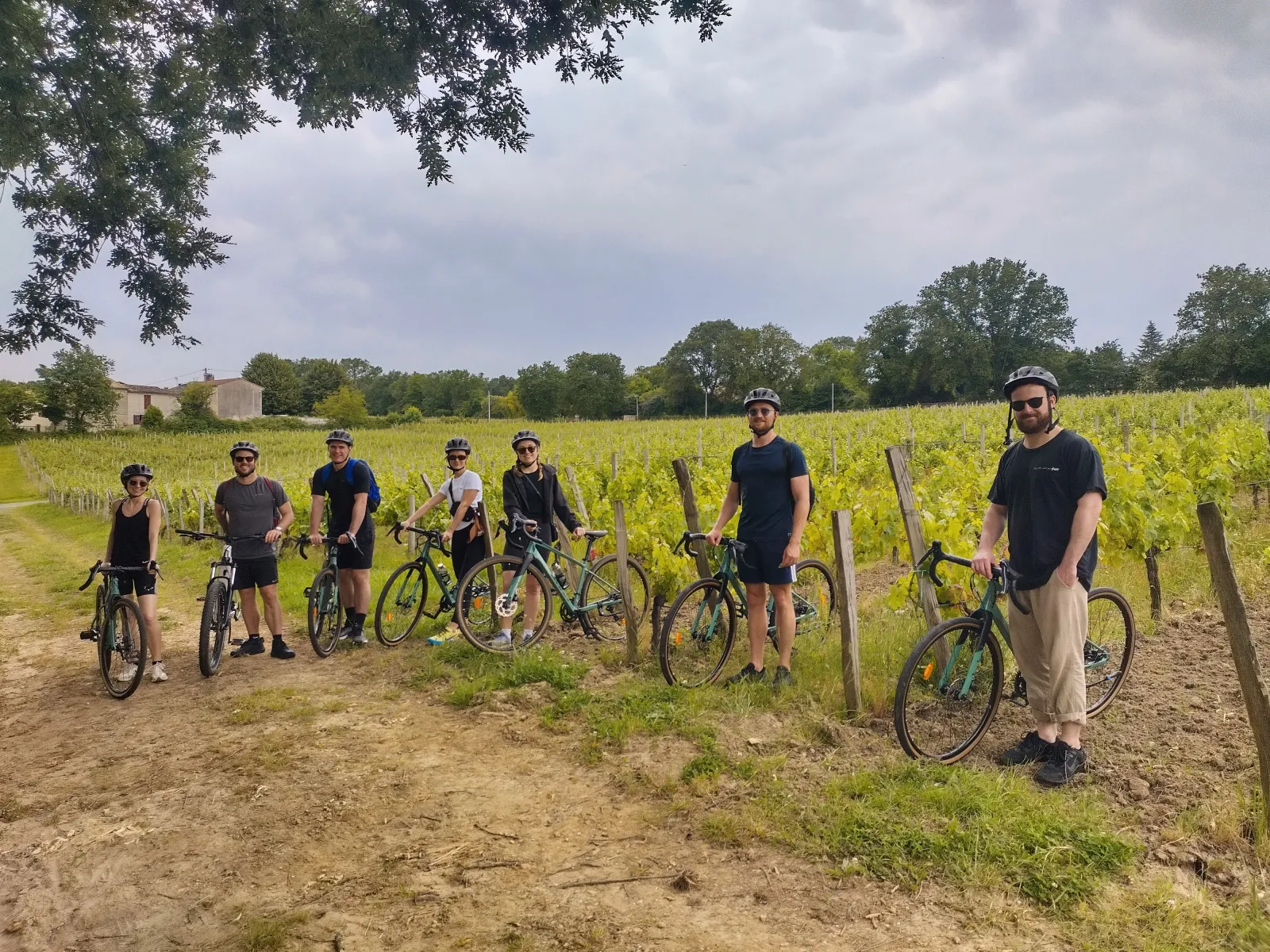 A group of cyclists with their gravel bikes posing on a dirt road between Bordeaux vineyards, with rows of green vines and stone buildings in the background.
