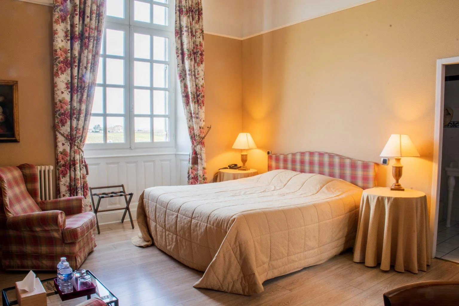 Elegant hotel bedroom with warm peach-yellow walls, double bed with white linens, bedside tables with lamps, red plaid armchair, large windows with floral curtains overlooking the Bordeaux countryside
