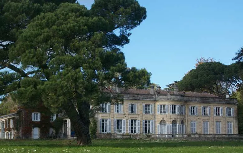 Château du Taillan in Bordeaux, a classical mansion with pale yellow facades and blue shutters, surrounded by large green trees and a lawn.