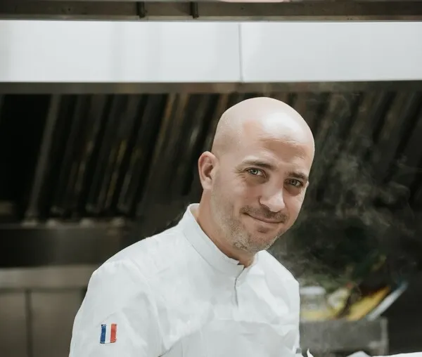 Smiling bald man wearing a white chef's jacket with French flag badge, posing inside Passage Secret restaurant in Bordeaux