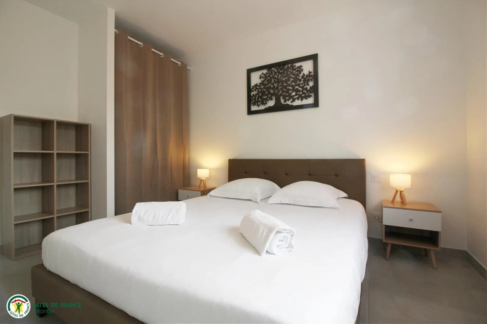 Modern bedroom in the Tanins guesthouse at Château Pierre de Montignac in Bordeaux, featuring a double bed with white linens, two nightstands with lamps, a brown upholstered headboard, brown curtains, a gray wall shelf, and tree silhouette wall art.