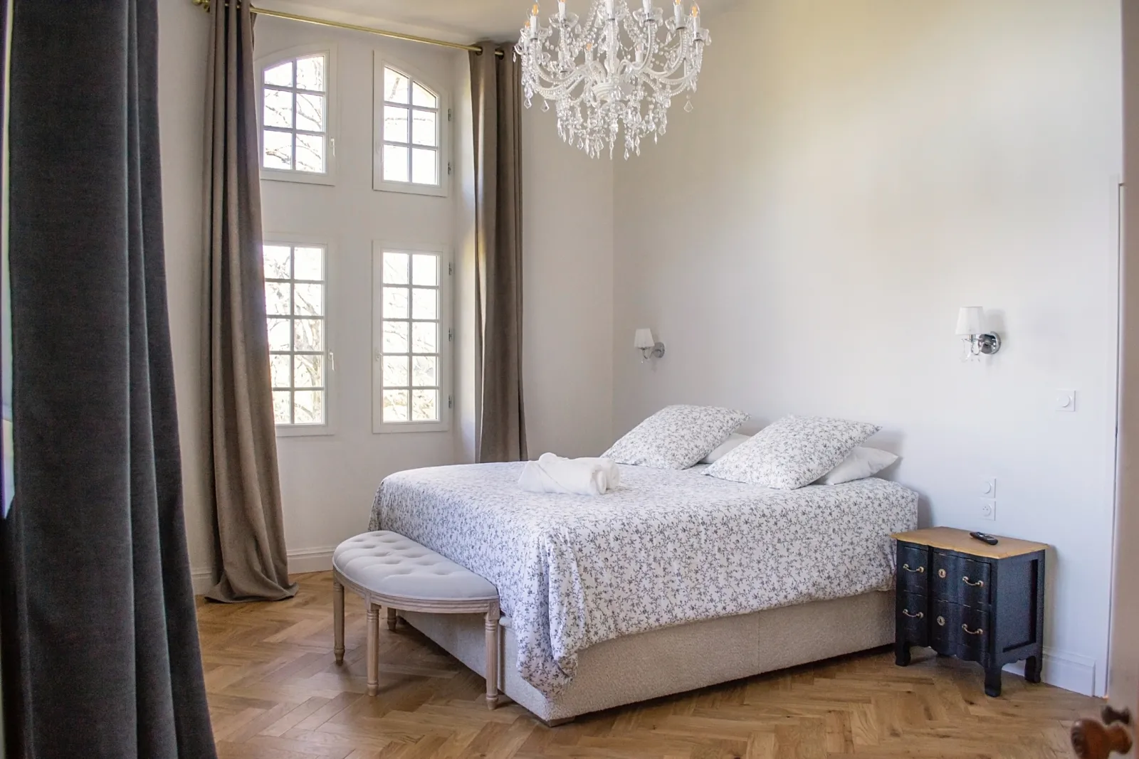 Elegant bedroom featuring a double bed with floral patterned linens, crystal chandelier, French doors overlooking gardens, herringbone flooring, and classic furnishings at La Réserve du Château de Roques in Bordeaux.