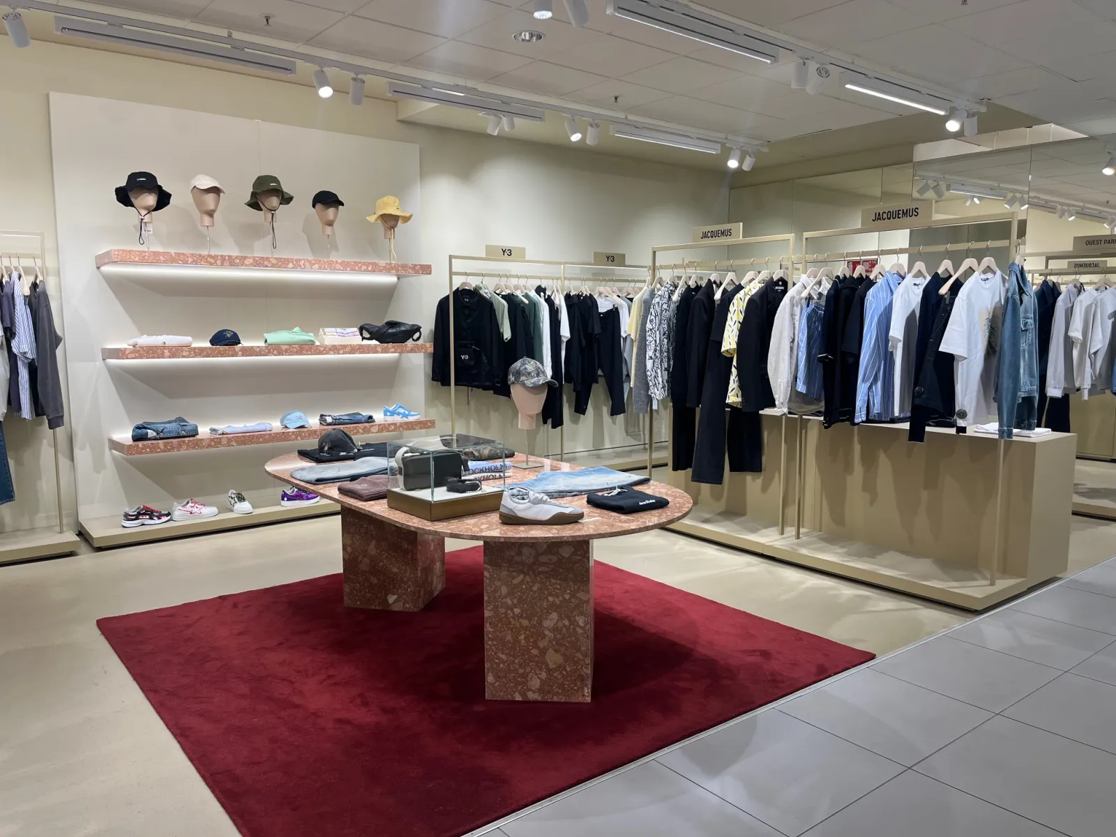 Interior of Galeries Lafayette Homme store in Bordeaux featuring a display area with hat shelves, central exhibition table on burgundy carpet, and clothing racks with classic garments