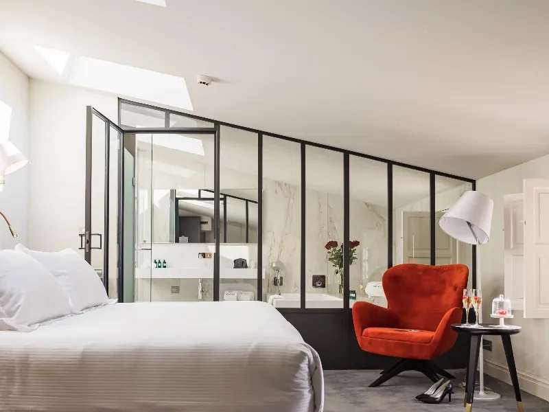 Modern hotel room at Villas Foch in Bordeaux featuring a white bed, black-framed glass bathroom, orange armchair, and contemporary furnishings