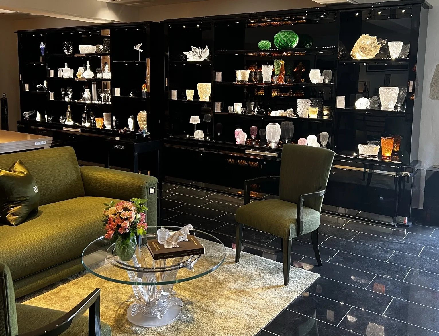 Luxury showroom at Château Lafaurie Peyraguey in Bordeaux featuring illuminated black display cabinets with premium glassware and vases, green velvet armchairs, and a round glass table with fresh flowers.